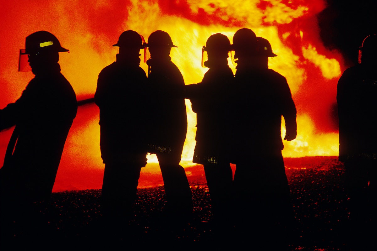 firefighters in full gear silhouetted in front of blazing fire
