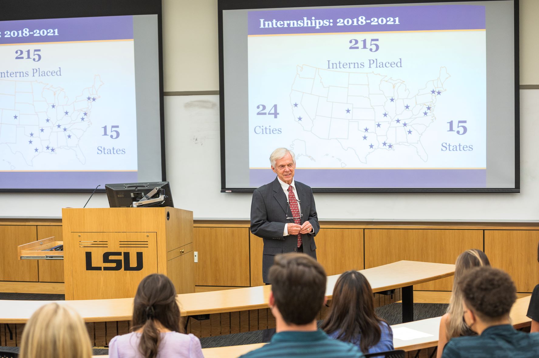 Professor in suit at the front of a classroom with screens behind him