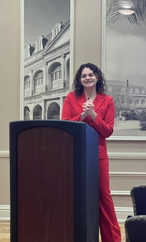 Madison Craig stands at the podium wearing a red suit to give her research presentation.