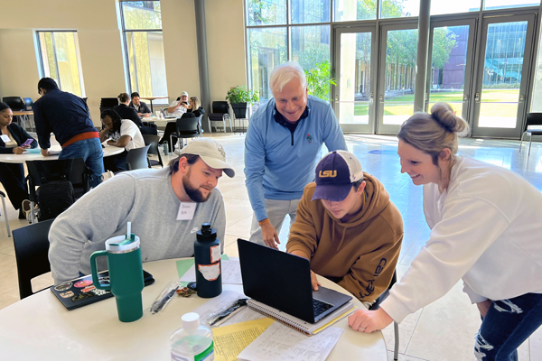A professional advises a group of restaurant management students as build a virtual cafe. The group chats excitedly and points at a laptop as they sit in the BEC Rotunda on a bright, sunny day.